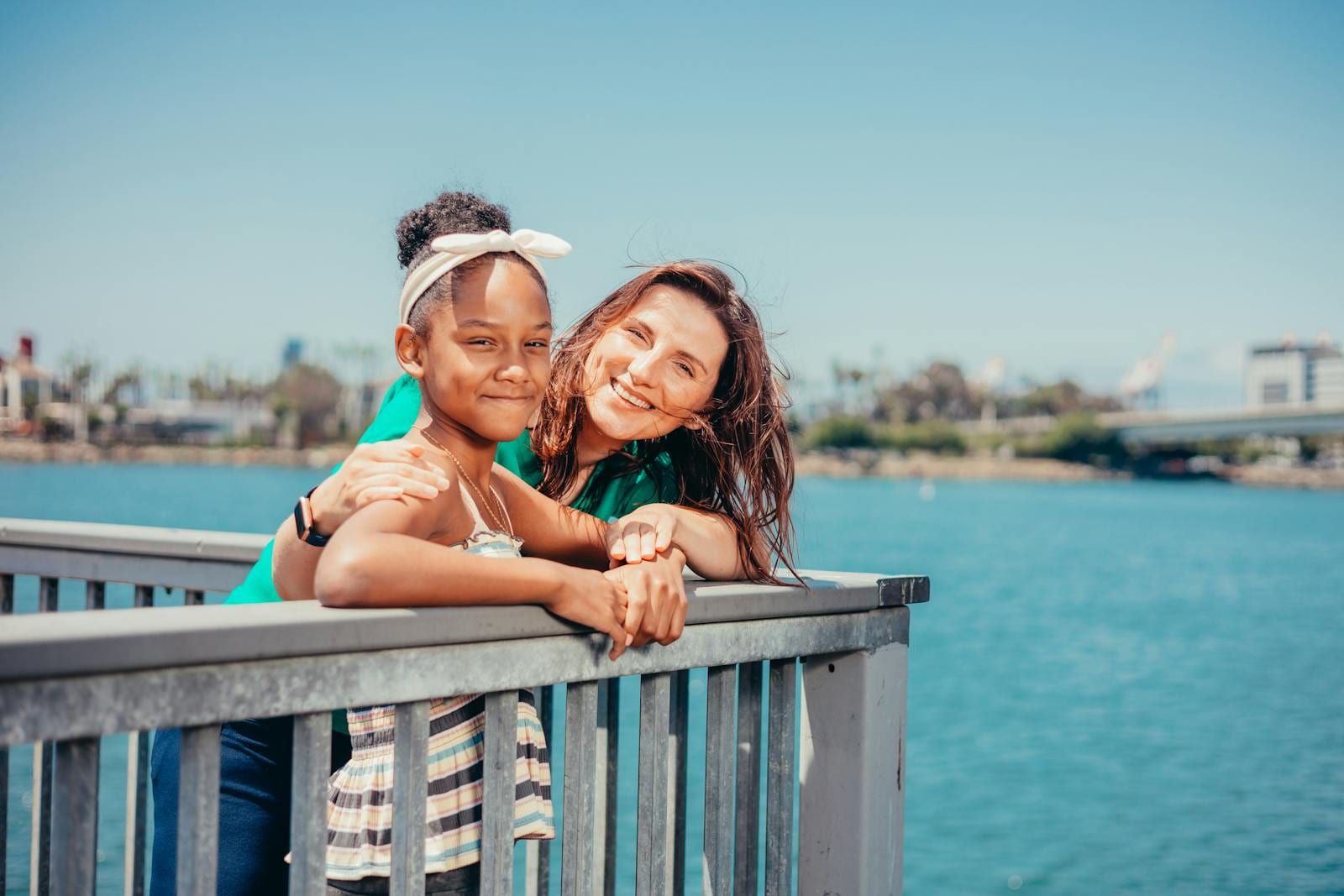 A smiling mother and daughter enjoying a sunny day by the waterfront with blue skies.